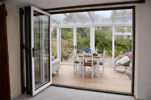 White internal bifold doors open into a bright conservatory dining area with a glass roof and garden view.